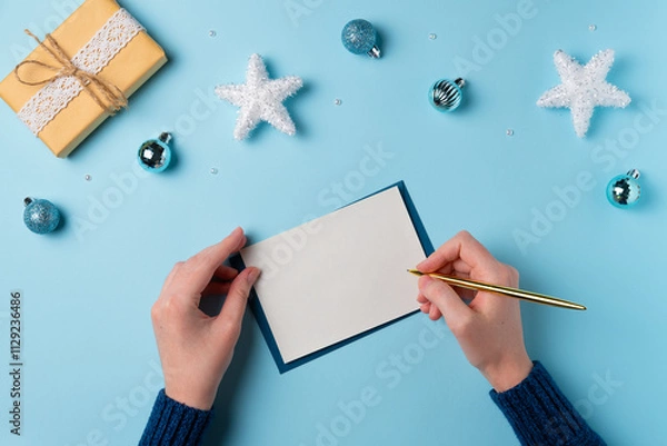 Fototapeta Female hand writing on a blank sheet of paper on a blue background. Christmas concept. Flat lay, top view