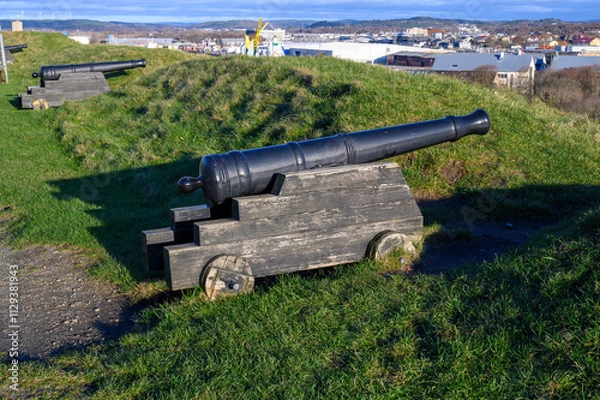 Obraz A historic cannon is positioned on grassy terrain, providing a stunning view of the city skyline under a clear blue sky. The scene captures a mix of nature and urban landscape