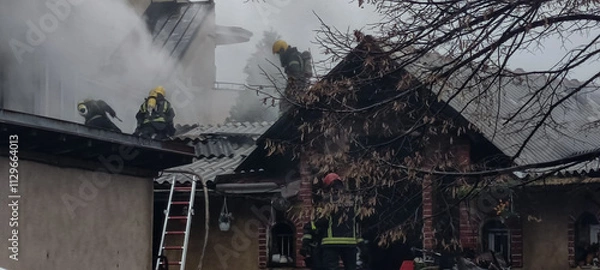 Obraz Firefighters climbing a ladder to the roof of a house, smoke coming out from behind and around it