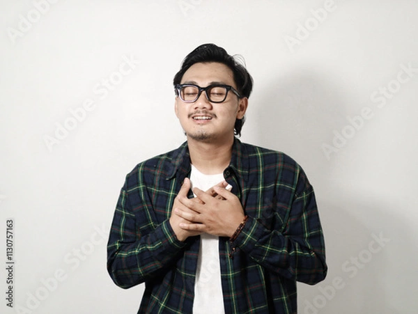 Obraz Portrait of smiling pleasant Young Asian man in plaid shirt looking camera and holding hands on his chest, heart isolated on white background