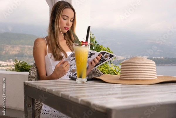 Fototapeta A woman is sitting at a table with a glass of orange juice and a book