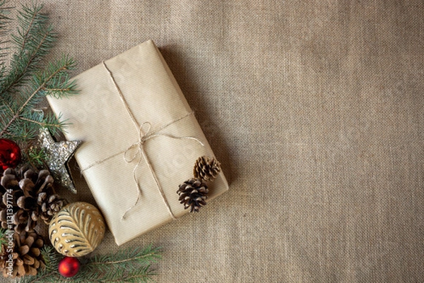 Obraz A gift wrapped in paper is placed on a burlap background, surrounded by Christmas decorations. Copy space. Vintage style.