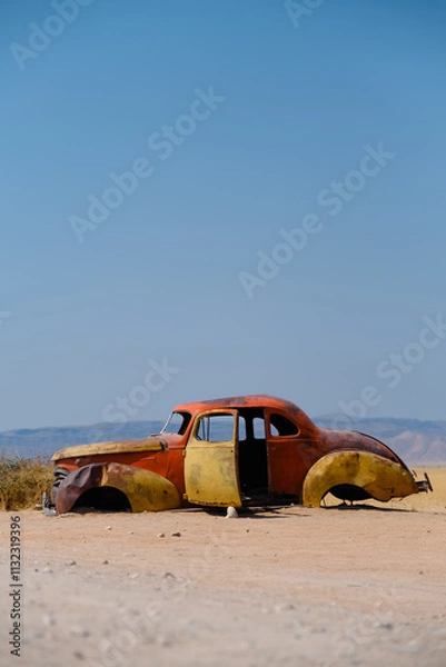 Obraz Vintage car abandoned in Solitaire, Namibia. Its rusty yellow and orange hues contrast with the arid sands and blue sky, reflecting nostalgia and the passage of time
