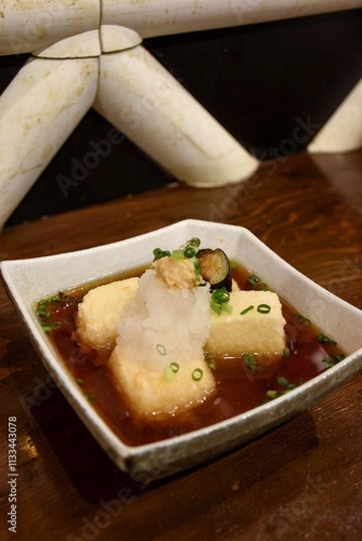 Fototapeta Crispy agedashi tofu cubes sit in a savory broth, topped with grated radish, chopped green onions, and a touch of ginger. Served in a square dish, the flavors reflect traditional Japanese cuisine.