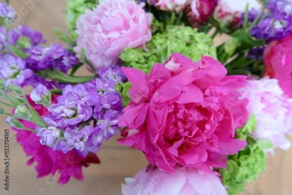 Fototapeta A vibrant floral arrangement featuring bold pink peonies, delicate purple blossoms, and fresh green accents, creating a lively and colorful composition with a softly blurred background.
