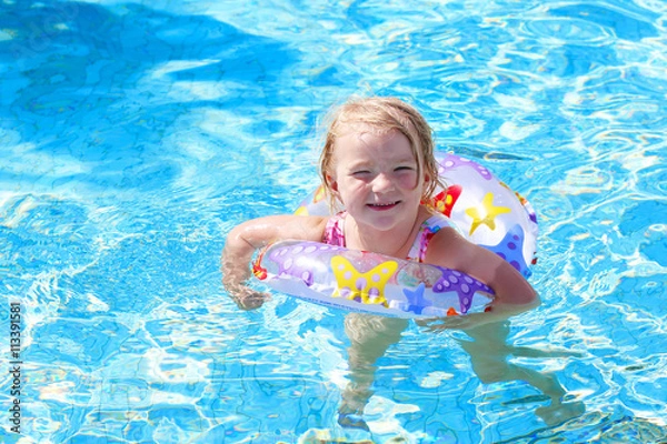Fototapeta Adorable happy little child, curly toddler girl in swimming suit having fun relaxing and floating on an inflatable toy ring in a pool on sunny day during summer vacation in resort
