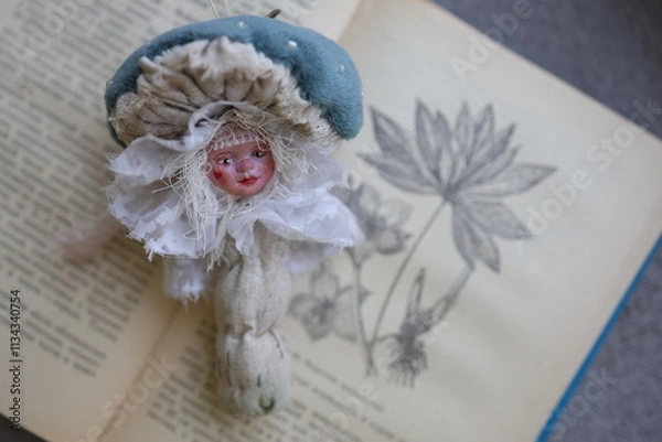 Fototapeta Unique doll resembling a mushroom placed on a vintage book with floral illustrations at home. A whimsical doll shaped like a mushroom with delicate features rests atop an open vintage book