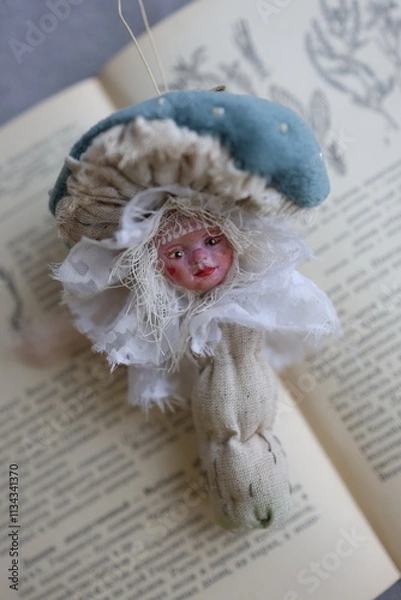 Fototapeta Unique doll resembling a mushroom placed on a vintage book with floral illustrations at home. A whimsical doll shaped like a mushroom with delicate features rests atop an open vintage book