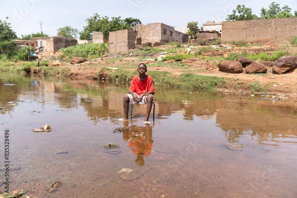 Obraz Young African boy sitting on a chair in the middle of a flooded area symbolizing the impact of climate change on life in poor developing countries