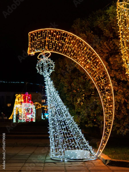Fototapeta Colorful holiday lights create a vibrant display in a park at night. A white tree structure stands next to a large golden arch, adding to the festive atmosphere that celebrates the season.