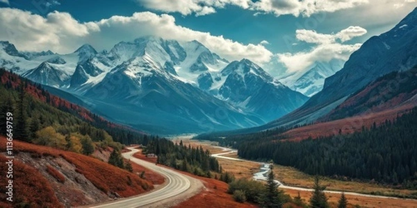 Fototapeta A winding asphalt road snakes through a valley, flanked by vibrant autumn foliage and towering snow-capped mountains under a sky of fluffy white clouds.