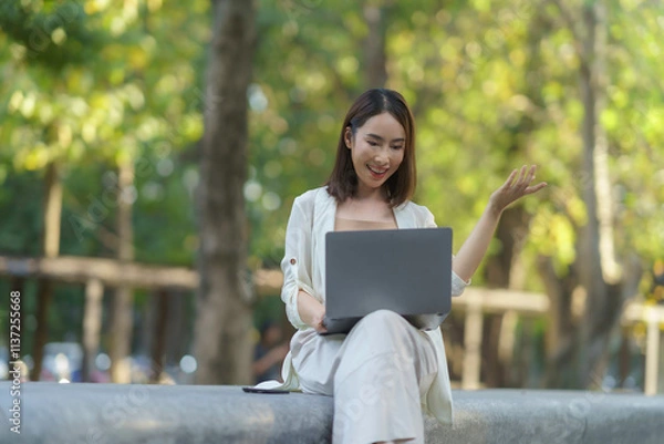 Fototapeta Asian businesswoman using laptop for video conference, working remotely in a park, enjoying work life balance