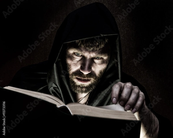 Fototapeta necromancer casts spells from thick ancient book by candlelight on a dark background