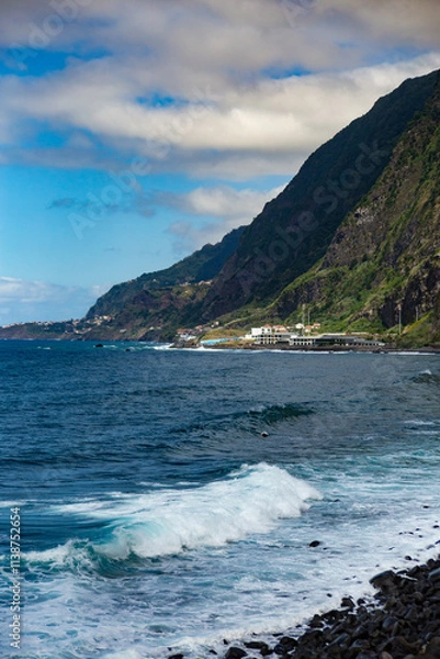 Fototapeta Powerful waves crashing against Madeira’s rocky shoreline, with steep green cliffs and the vast Atlantic Ocean under a partly cloudy sky.