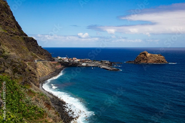 Fototapeta Powerful waves crashing against Madeira’s rocky shoreline, with steep green cliffs and the vast Atlantic Ocean under a partly cloudy sky.