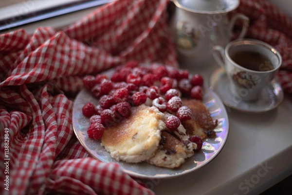 Obraz Golden-brown cottage cheese pancakes served with fresh raspberries, lightly dusted with powdered sugar. The plate is accompanied by a vintage teapot, a cup of tea, and a red gingham cloth, creating a 