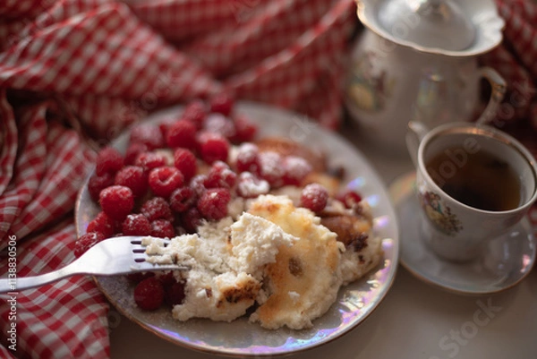 Obraz Golden-brown cottage cheese pancakes served with fresh raspberries, lightly dusted with powdered sugar. The plate is accompanied by a vintage teapot, a cup of tea, and a red gingham cloth, creating a 
