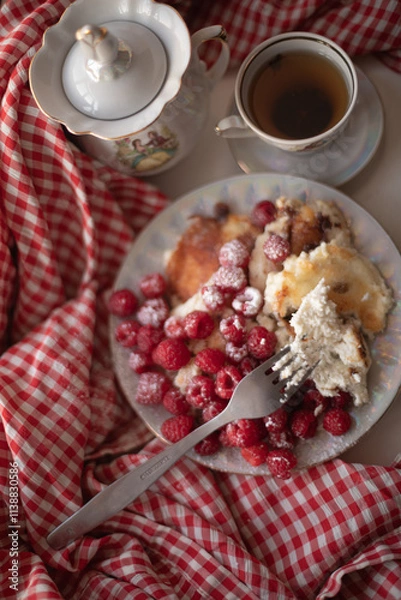 Obraz Golden-brown cottage cheese pancakes served with fresh raspberries, lightly dusted with powdered sugar. The plate is accompanied by a vintage teapot, a cup of tea, and a red gingham cloth, creating a 