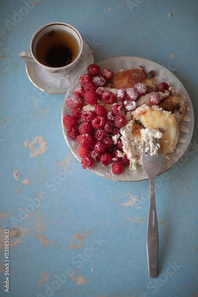 Obraz Golden-brown cottage cheese pancakes served with fresh raspberries, lightly dusted with powdered sugar. The plate is accompanied by a vintage teapot, a cup of tea, and a red gingham cloth, creating a 