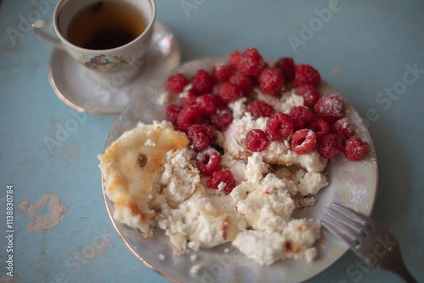 Obraz Golden-brown cottage cheese pancakes served with fresh raspberries, lightly dusted with powdered sugar. The plate is accompanied by a vintage teapot, a cup of tea, and a red gingham cloth, creating a 