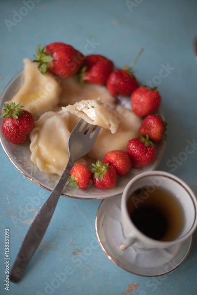 Obraz A plate of sweet dumplings with strawberries on a light blue table, with a bite taken out to reveal the creamy filling. A cup of tea sits nearby, completing the relaxing scene.
