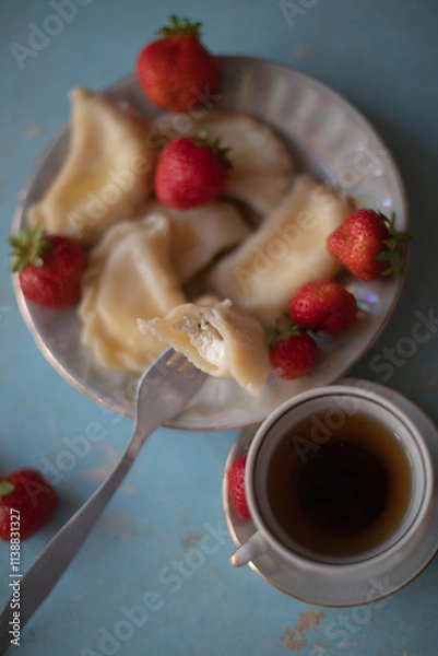 Obraz A plate of sweet dumplings with strawberries on a light blue table, with a bite taken out to reveal the creamy filling. A cup of tea sits nearby, completing the relaxing scene.
