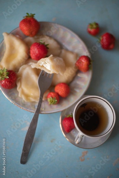 Obraz A plate of sweet dumplings with strawberries on a light blue table, with a bite taken out to reveal the creamy filling. A cup of tea sits nearby, completing the relaxing scene.
