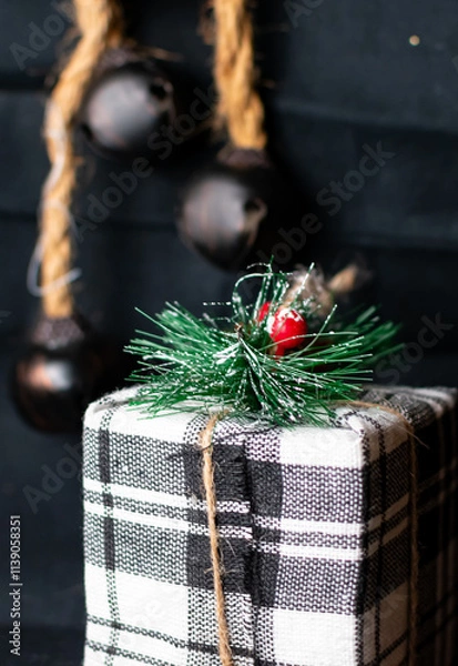 Fototapeta A Christmas tree ornament that is shaped like a present with blurred jingle bells in the distant background. 