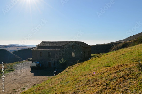 Fototapeta A building situated on a hill, surrounded by grass, with a clear sky and mountainous background reflecting a rural landscape