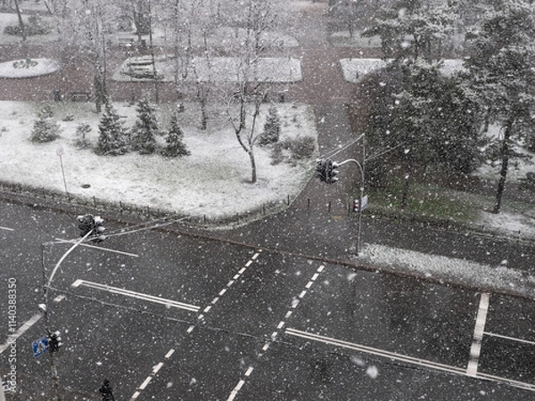 Fototapeta View from the window over heavy fluffy snow falling over the pedestrian crossing with red traffic light and urban park with a lonely person walking. Snowy winter day.