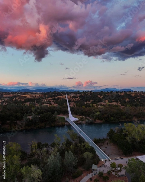 Fototapeta Stunning aerial view of the Sundial Bridge at dusk, with vibrant pink and purple clouds reflecting the fading light over the river and surrounding landscape.