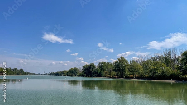 Fototapeta Wide view of a serene lake reflecting the sky, surrounded by lush trees under a bright blue sky, ideal for nature, relaxation, and eco-tourism themes.