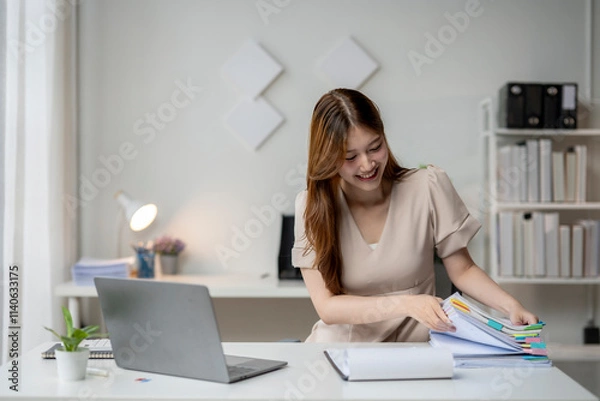 Fototapeta A woman is sitting at a desk with a laptop and a stack of papers