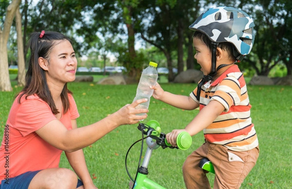 Fototapeta Mother giving the drinking water to boy on the bicycle in garden