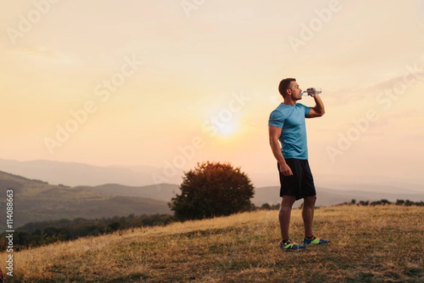 Fototapeta Athletic man in his 30s resting and drinking water from a bottle while sitting down outdoor