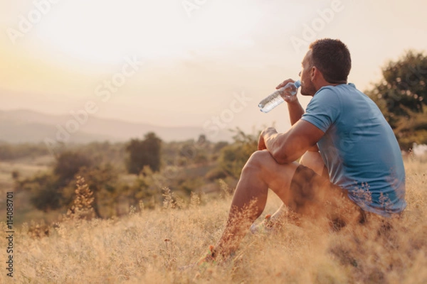 Fototapeta Athletic man in his 30s resting and drinking water from a bottle while sitting down outdoor