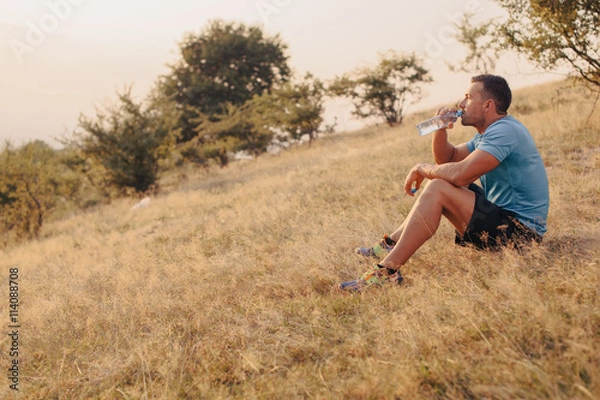 Fototapeta Athletic man in his 30s resting and drinking water from a bottle while sitting down outdoor