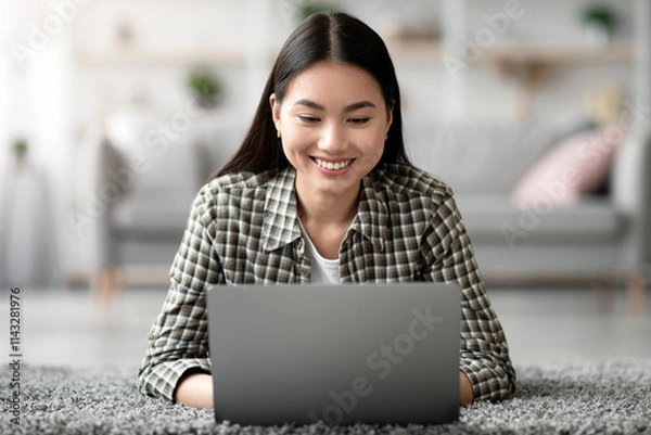 Fototapeta Relaxed millennial asian woman looking for job opportunities online, laying on carpet amd using modern laptop at home, smiling and typing on notebook keyboard, chinese lady working remotely, closeup