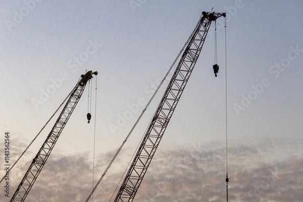 Fototapeta Dec 16 2024,Hong Kong.A series of construction cranes rise against a colorful sunset, casting long shadows over a busy construction site