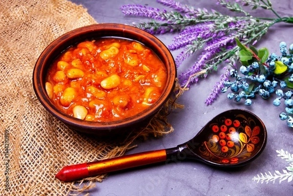 Fototapeta Bean soup, beans in tomato in a clay plate, wooden spoon, burlap, berries and herbs on a purple background