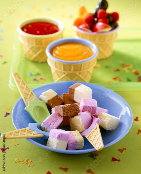 Fototapeta Home made ice cream for children birthday party. A plate features an assortment of colorful ice cream squares alongside bowls of syrup and fruit.