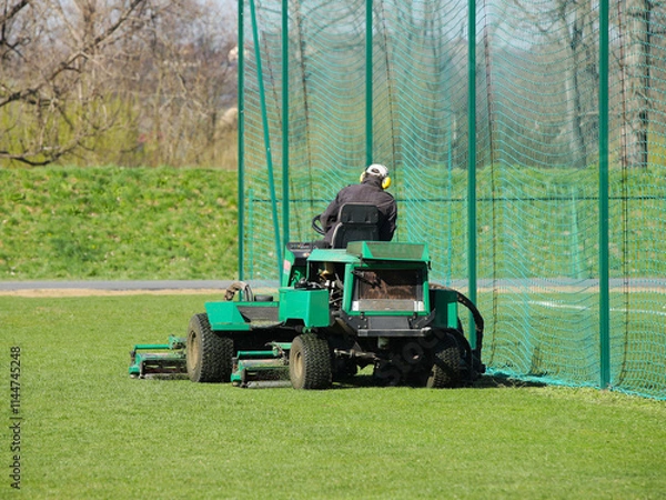 Fototapeta Worker on a large green lawn mower mows the grass on the football field. Landscape design and maintenance of green areas of the sports complex. Making grass stadium before the match. Human labor
