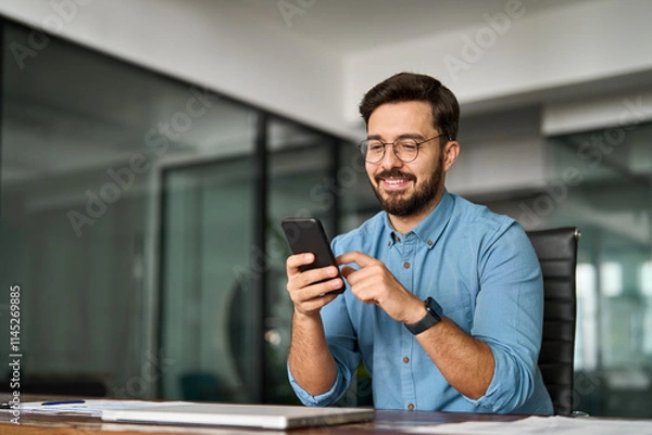 Fototapeta Busy young Latin business man holding smartphone working in office. Professional happy businessman entrepreneur or manager using financial banking apps making easy payments on cell phone at work.