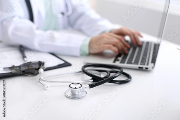 Fototapeta Close-up of stethoscope is lying on the table near female doctor typing on laptop computer