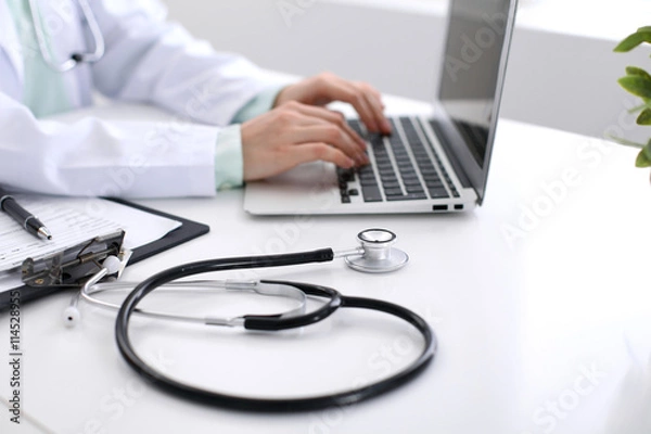 Fototapeta Close-up of stethoscope is lying on the table near female doctor typing on laptop computer