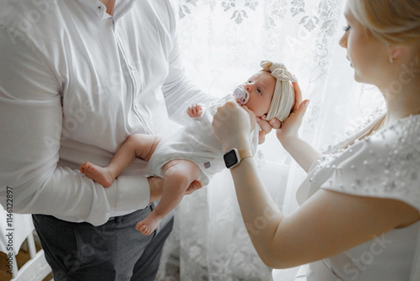 Fototapeta A man and a woman are holding a baby. The baby is wearing a white hat and a pacifier. The scene is warm and loving, with the family bonding over the new addition to their family