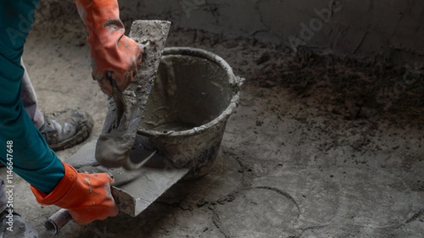 Obraz A worker uses a trowel to scoop up cement from a black plastic bucket while plastering a house wall. Concept of working in a construction site