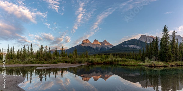 Fototapeta Three Sisters in Canmore seen at golden hour, sunset on blue sky day, afternoon with calm, peaceful reflection in water below famous, tourist, tourism mountains, area summer.	