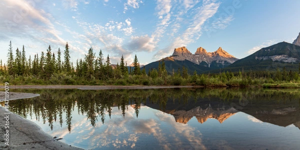 Fototapeta Three Sisters in Canmore seen at golden hour, sunset on blue sky day, afternoon with calm, peaceful reflection in water below famous, tourist, tourism mountains, area summer.	