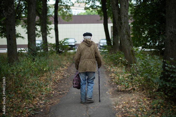 Fototapeta Old man walks along path in park. Poor old man walking down street. Pensioner with walking stick.